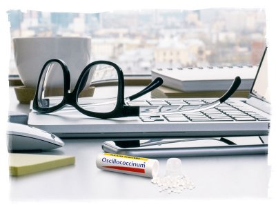 Tube of Oscillo with pellets spilled out on work desk next to phone, sticky pad, mouse, laptop, eyeglasses, books, and coffee mug, in front of window with city in background